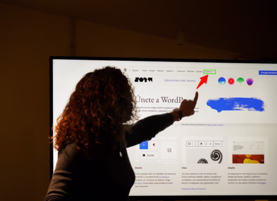 A person with curly hair is silhouetted against a large computer screen, pointing at a section of a webpage. The screen displays a website in Spanish, featuring the WordPress logo and navigation menu, with a red arrow pointing to the “Colabora” menu item.